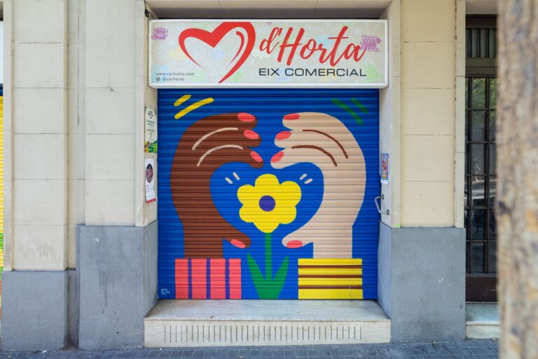 Persiana del eje comercial Cor d'Horta pintada por Emily Eldridge con dos manos que sostienen una flor amarilla dentro de un corazón — Persianas que hacen barrio, Cor d'Horta, Barcelona, Rebobinart 2026