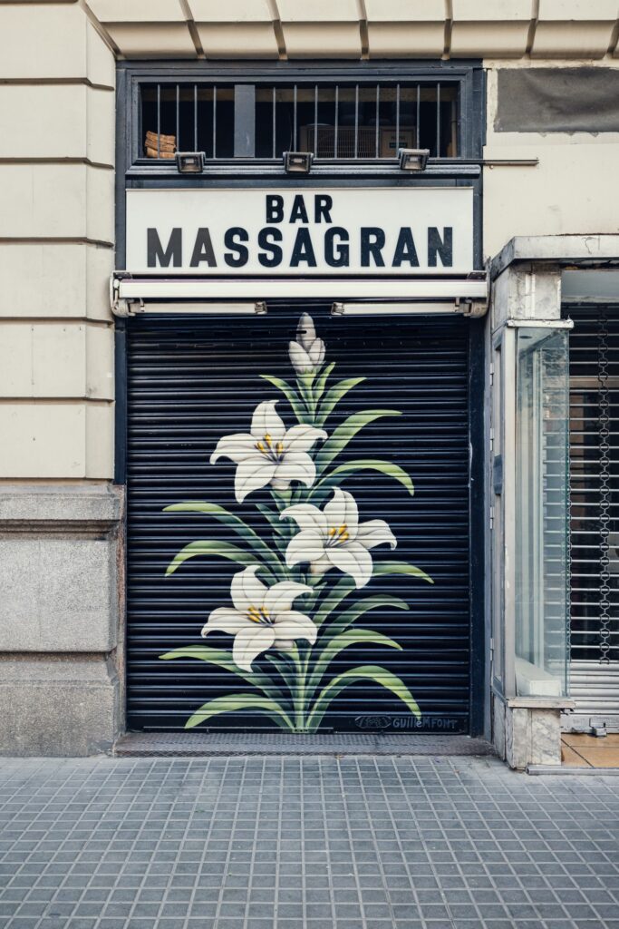 persianas-que-hacen-barrio-bar-massagran-guillem-font Persiana del Bar Massagran pintada por Guillem Font con tres lirios blancos, los de la bandera de la Vila de Gràcia, sobre fondo negro — Persianes que fan barri, Gràcia, Barcelona, Rebobinart 2026