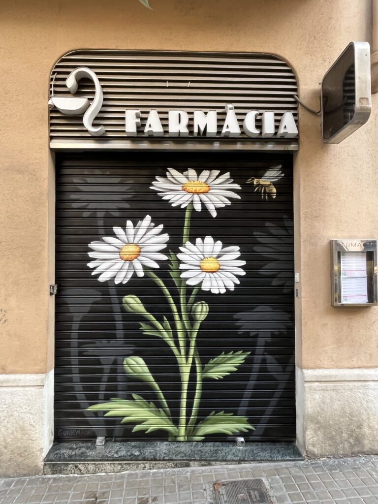 persianas-que-hacen-barrio-farmacia-adela-garcia-lopez-guillem-font Persiana de la Farmàcia Adela Garcia López pintada por Guillem Font con margaritas y una abeja sobre fondo negro — Persianes que fan barri, Gràcia, Barcelona, Rebobinart 2026