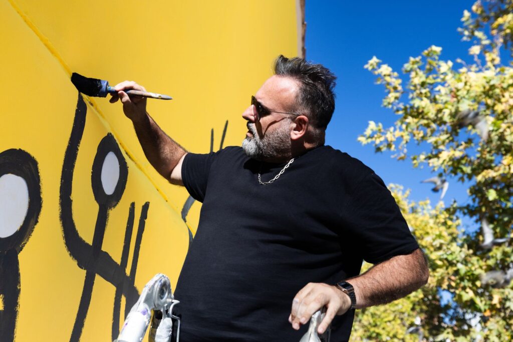 Javirroyo amb el pinzell a la mà davant dels personatges del mural, artista d'humor gràfic, CASA NOSTRA, Barcelona, Rebobinart 2026