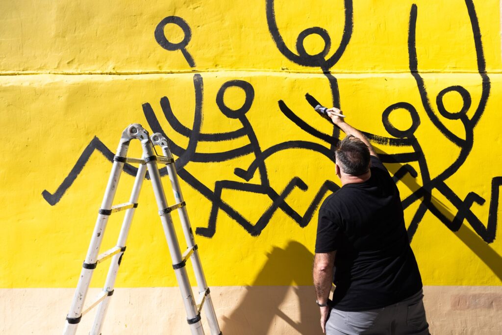 Javirroyo pintant la filera de personatges negres sobre fons groc, mural d'humor gràfic CASA NOSTRA, Barcelona, Rebobinart 2026