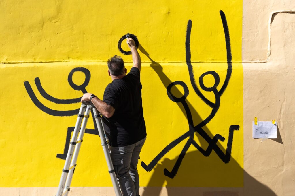 Javirroyo pinta en negre els personatges que corren, enfilat a una escala, mural CASA NOSTRA, Barcelona, Rebobinart 2026