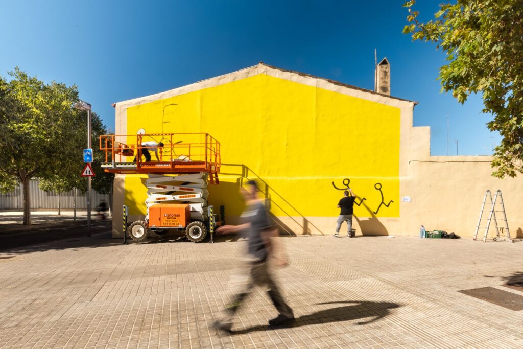 Javirroyo comença a pintar el mur groc del carrer Trinxant amb la plataforma elevadora al costat, mural CASA NOSTRA, Barcelona, Rebobinart 2026