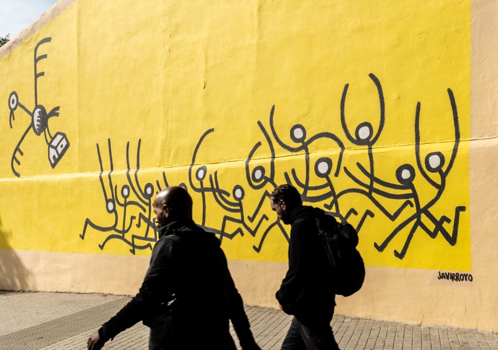 Dos vianants a contrallum passen davant del mural groc de Javirroyo, CASA NOSTRA, Barcelona, Rebobinart 2026
