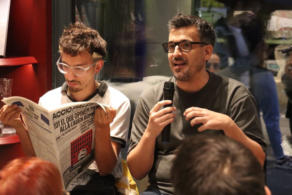 diari-accio-palestina-the-posttraumatic-presentacio Ponent de The PostTraumatic parlant mentre un altre llegeix el diari Acció Palestina, Llibreria Finestres Palestina, Barcelona, Rebobinart 2026