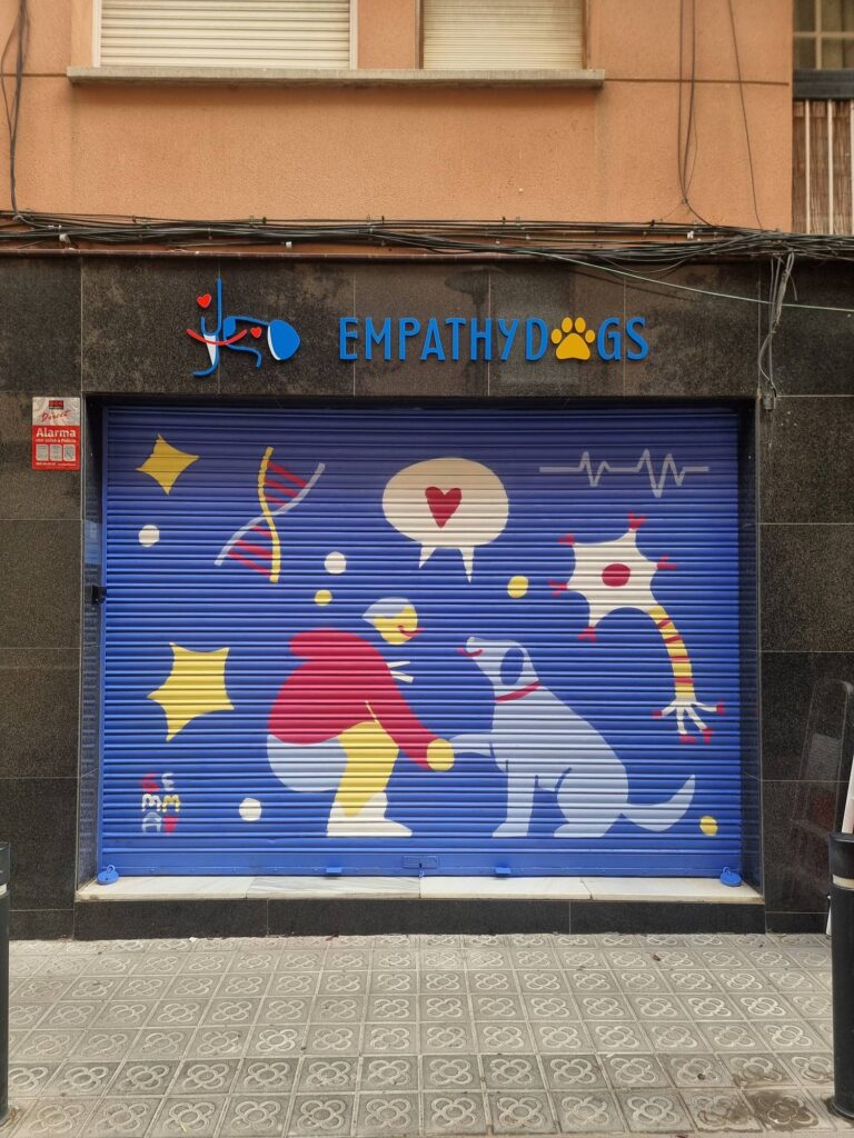 Persiana de Empathydogs pintada por Gemma Fontanals con una figura agachada ante un perro blanco, un corazón en un bocadillo, una hélice de ADN y una línea de electrocardiograma, sobre fondo azul — Persianes que fan barri, Sant Andreu, Barcelona, Rebobinart 2026