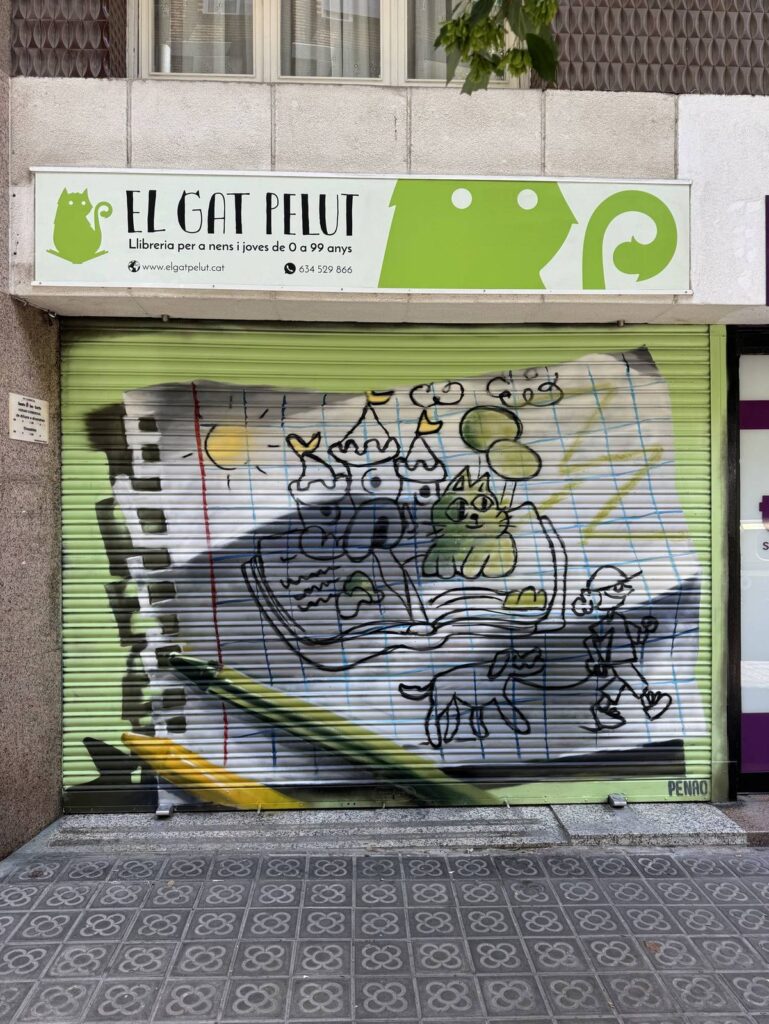 Persiana llibreria el gat pelut — Penao, Les Corts Persiana de la Llibreria El Gat Pelut pintada per Penao amb un full de quadern on surt un llibre obert del qual neixen un castell, un gat i personatges dibuixats amb línia infantil, i un llapis groc i verd en relleu, sobre fons verd — Persianes que fan barri, Les Corts, Barcelona, Rebobinart 2026