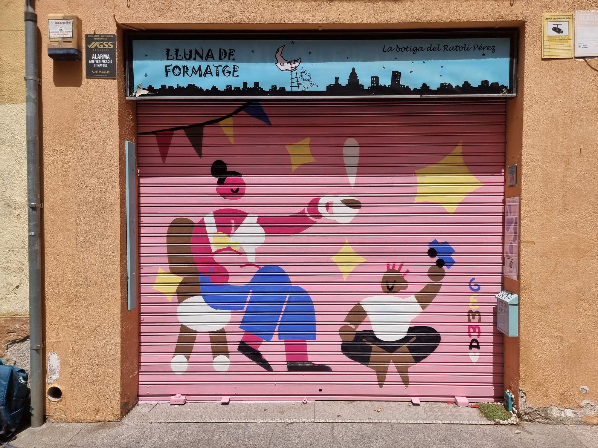 Persiana de Lluna de Formatge pintada por Gemma Fontanals con una figura adulta dando un diente a una criatura con corona, bajo banderines y estrellas amarillas, sobre fondo rosa — Persianes que fan barri, Sant Andreu, Barcelona, Rebobinart 2026