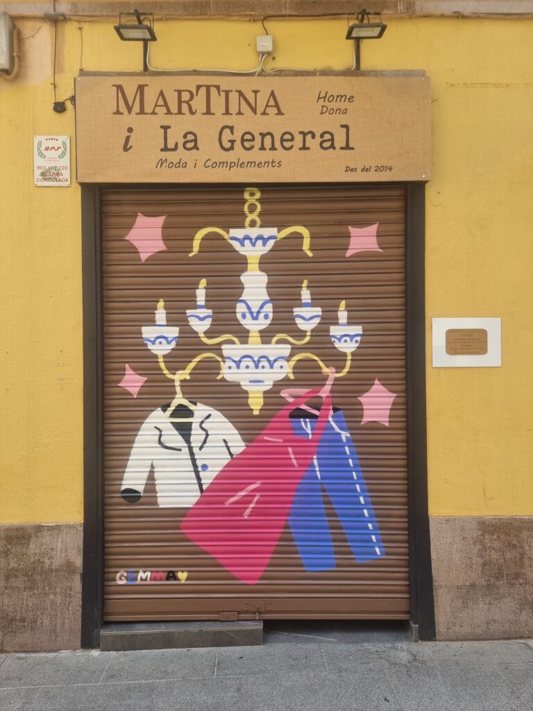 Persiana de Martina i la General pintada por Gemma Fontanals con una lámpara de araña blanca y azul sobre una americana clara y un abrigo rojo, sobre fondo marrón — Persianes que fan barri, Sant Andreu, Barcelona, Rebobinart 2026