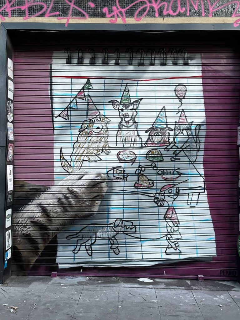 Persiana terranova — Penao, Les Corts Persiana de Terranova pintada per Penao amb una festa de gossos i gats amb barrets de festa dibuixada sobre un full de quadern i una pota de gat realista que s'hi recolza, sobre fons lila — Persianes que fan barri, Les Corts, Barcelona, Rebobinart 2026